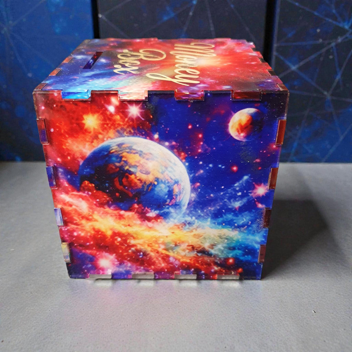 Front angled view of space themed money box with engraved “Money Box” lettering on lid and colourful planet artwork on the sides. Decorative acrylic savings box designed for kids bedroom or gift.