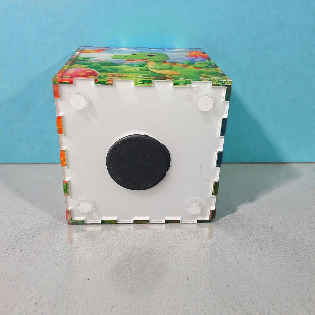 Bottom view of dinosaur themed acrylic money box showing removable white base with black rubber stopper for easy coin access. Durable construction with interlocking acrylic panels.