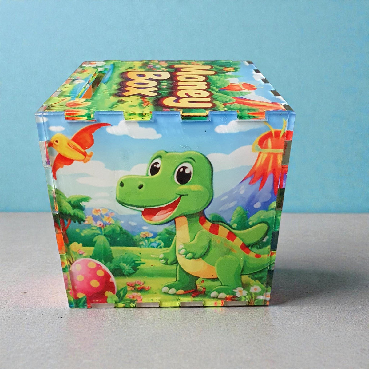 Green cartoon dinosaur money box with volcano and jungle background design. Colourful children’s savings box with coin slot on lid, suitable for bedroom décor or gift.
