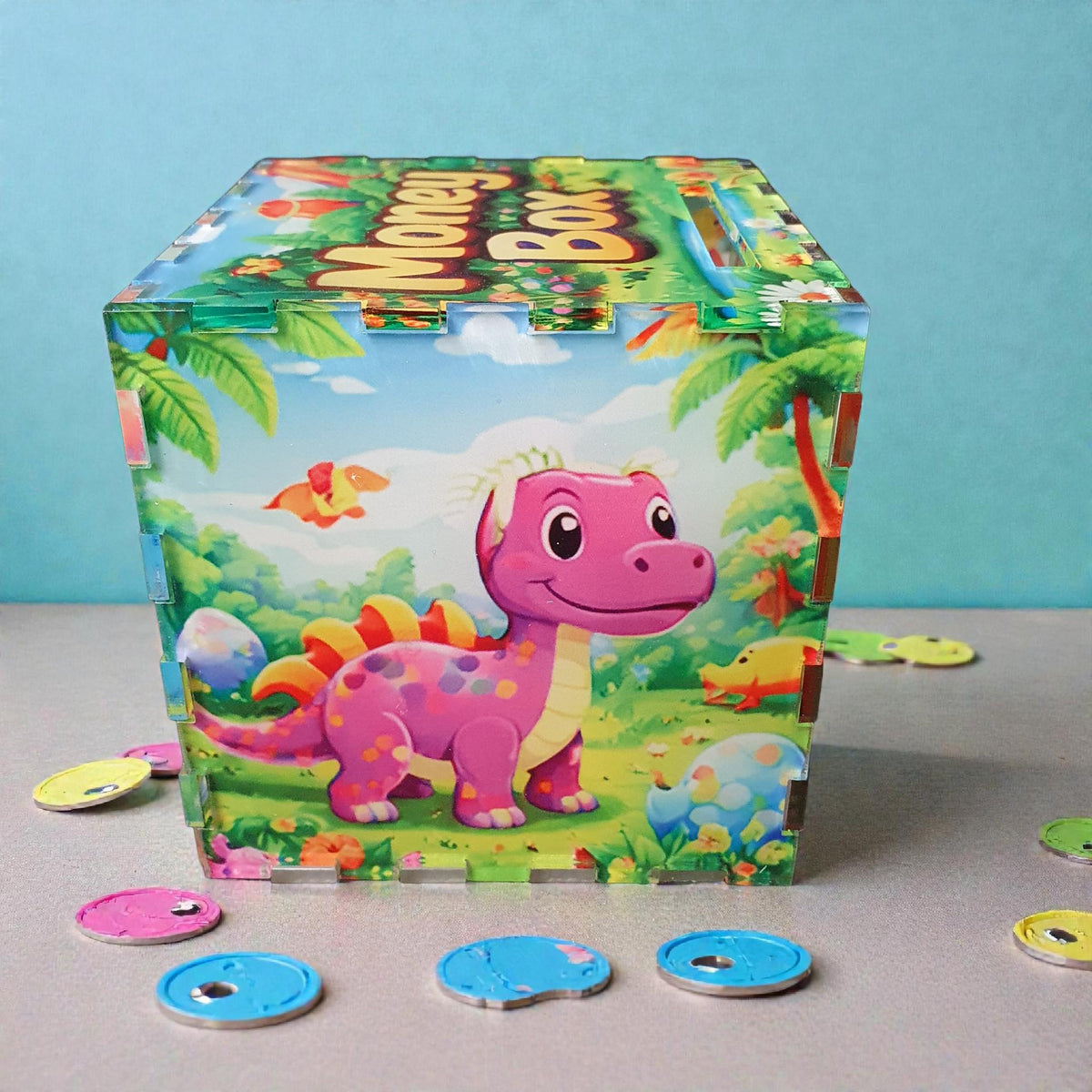 Side view of dinosaur money box featuring a purple cartoon dinosaur with bright eggs and tropical plants in the background. Acrylic children’s piggy bank with coin slot on top and colourful printed panels.