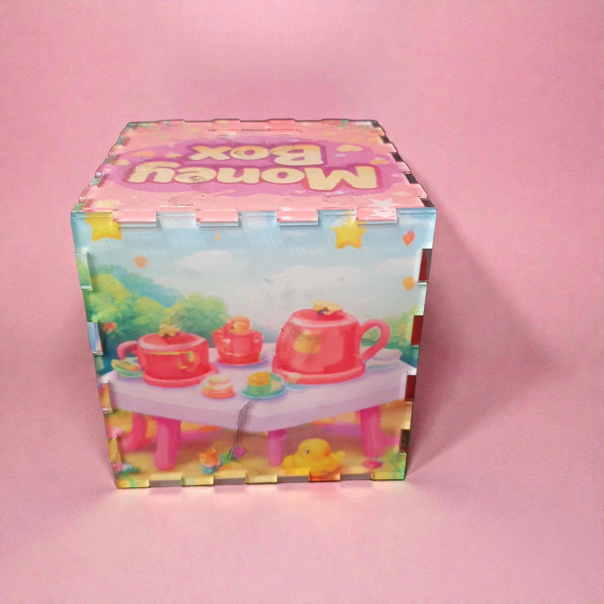 Opposite side of pink money box showing toy tea set with teapot, cups and small duck illustration. Cute pastel themed children’s savings box with colourful printed artwork.