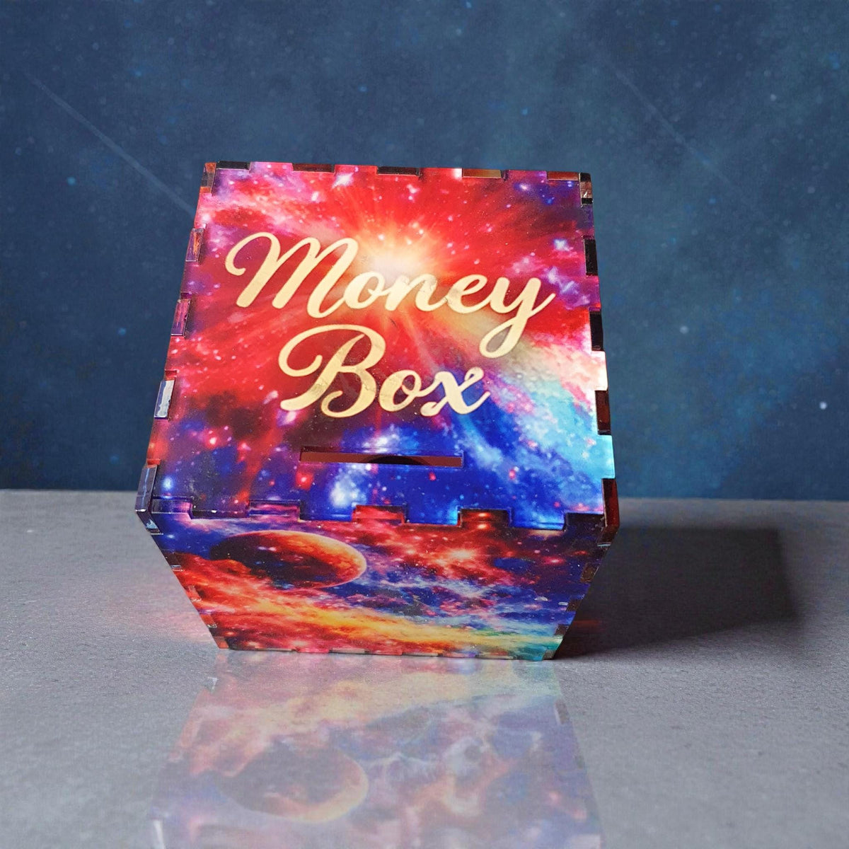 Galaxy themed acrylic money box with “Money Box” engraved on the lid, featuring vibrant space artwork in red, blue and purple tones. Decorative savings box displayed on a grey surface with a starry background, suitable for children or space lovers.