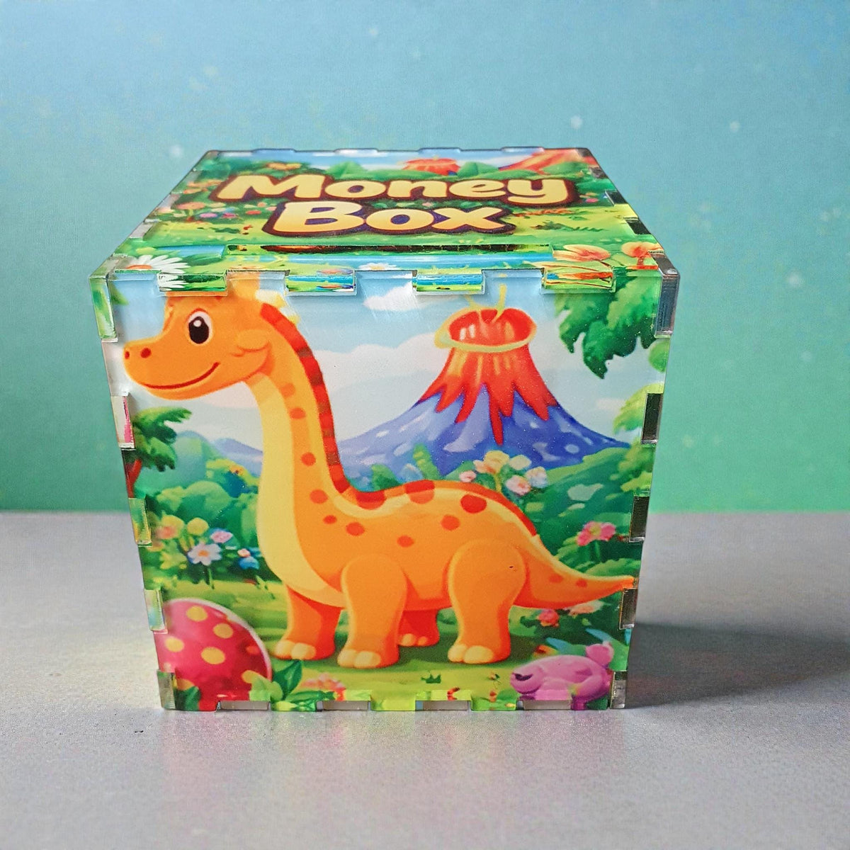 Children’s dinosaur themed acrylic money box with “Money Box” printed on the lid. The front features a smiling orange dinosaur in a colourful prehistoric landscape with volcano, mountains and flowers. Decorative kids savings box displayed on a neutral surface.