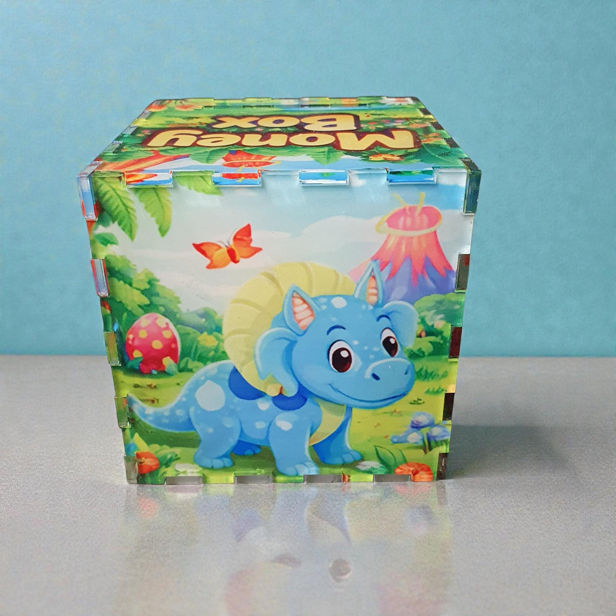 Front view of blue triceratops style dinosaur money box with volcano and jungle scene artwork. Kids savings box with vibrant prehistoric illustration and clear acrylic interlocking edges.