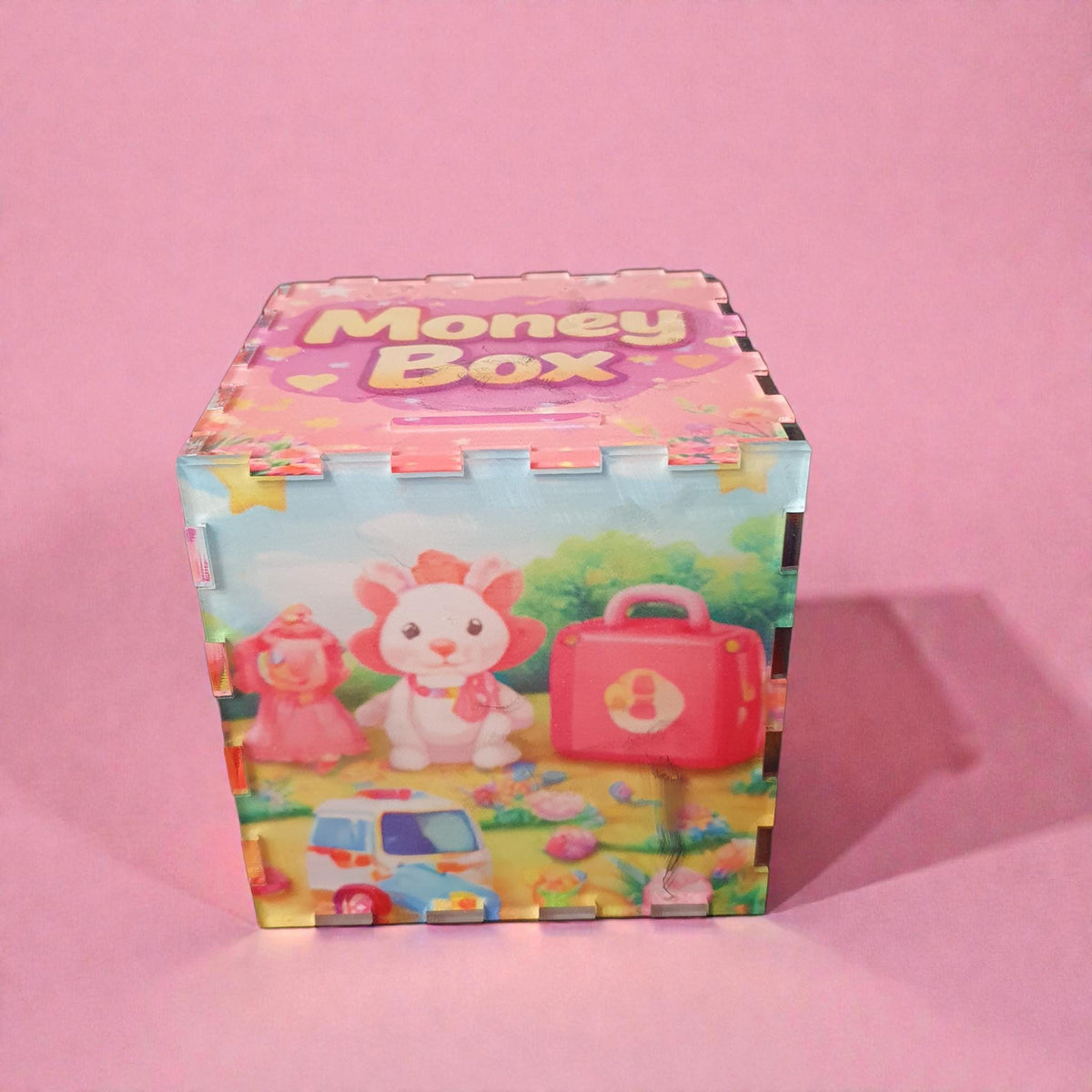 Front view of pink cartoon themed money box with bunny character, red suitcase and toy ambulance illustration. Acrylic children’s piggy bank with coin slot on top and colourful printed panels.