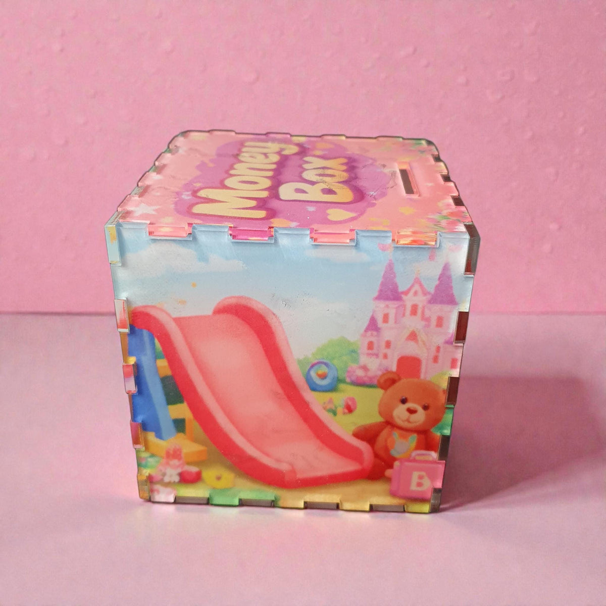 Side panel of pink children’s money box featuring playground slide, teddy bear and fairy-tale castle illustration. Bright pastel toy themed design printed on clear acrylic savings box.
