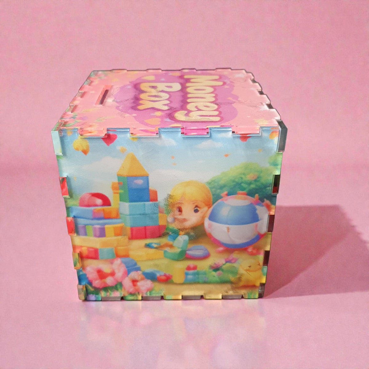 Another side view of pink kids money box featuring child playing with building blocks and ball in garden scene. Vibrant pastel toy design printed on acrylic piggy bank.