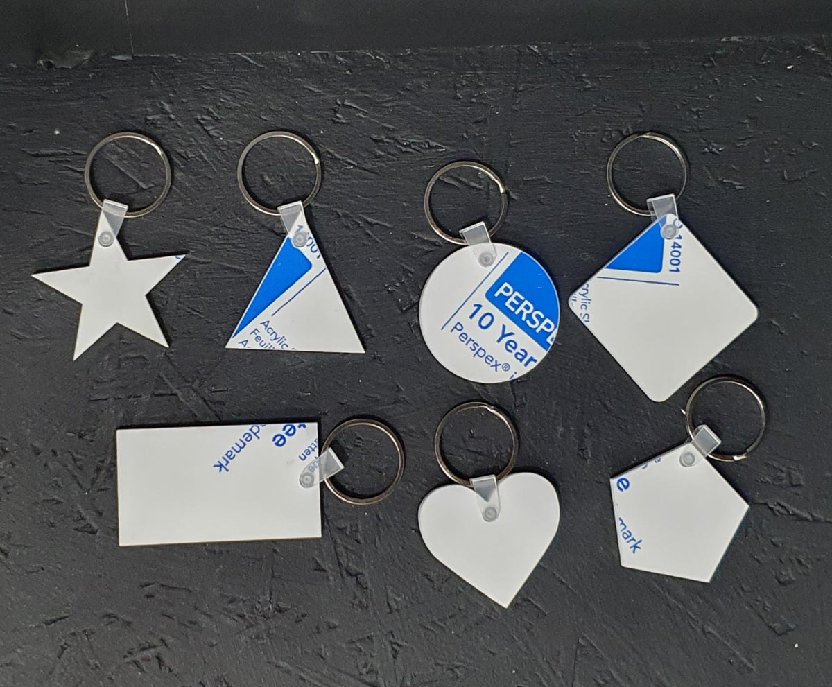 Overview image of assorted white cast acrylic keyring blanks including star, triangle, circle, square, rectangle, pentagon and heart shapes, each fitted with clear tag connector and 30mm metal split ring, displayed on dark background ready for personalisation.
