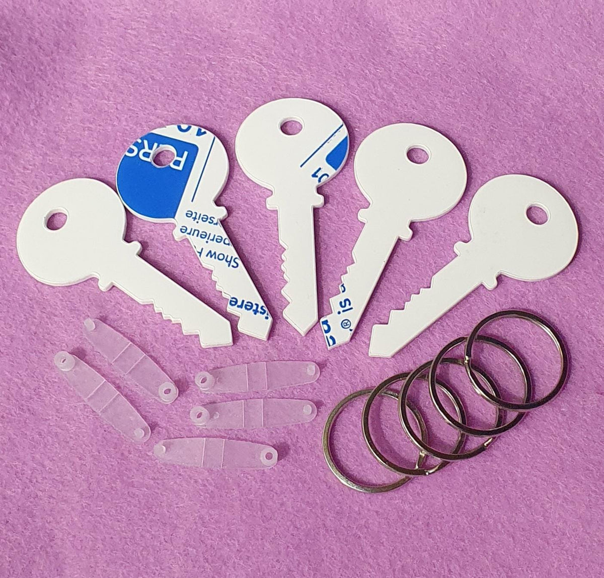 Set of 3mm acrylic key shaped keyring blanks measuring 7.5cm x 3.5cm displayed with included clear snap clips and metal split keyrings on pink background.