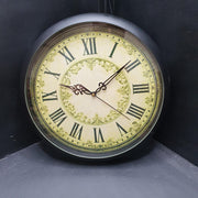 A circular antique clock face featuring Roman numerals. The clock has a cream-coloured background with black numerals and hands. It is framed with a gold-coloured border and decorative accents.