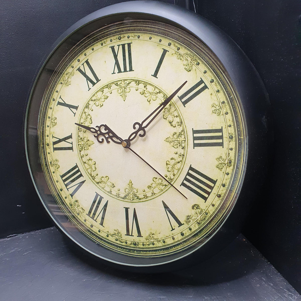 A circular antique clock face featuring Roman numerals. The clock has a cream-coloured background with black numerals and hands. It is framed with a gold-coloured border and decorative accents.