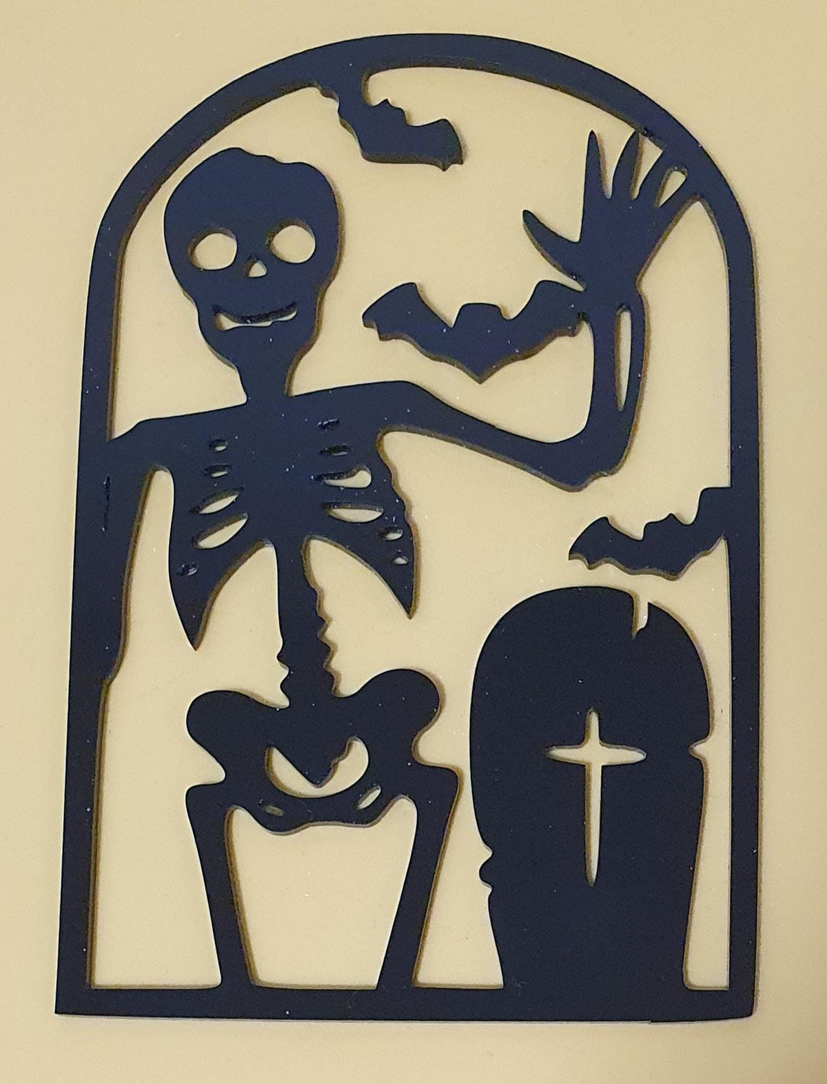 Black laser-cut acrylic Halloween decoration featuring a waving skeleton, flying bats, a spooky pumpkin and a cross gravestone in an arched frame.