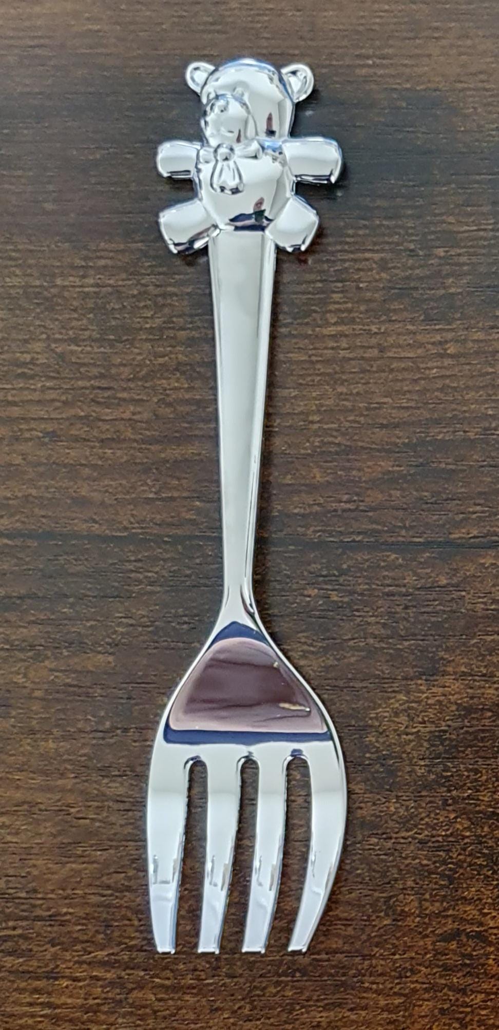 Silver plated fork on a wooden surface, featuring four rounded prongs and a teddy bear with a bow at the top of its handle.