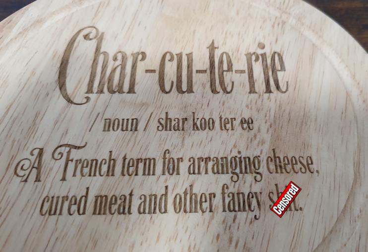 Round wooden cheese board with funny engraved charcuterie definition and four cheese tools stored neatly in the open base.