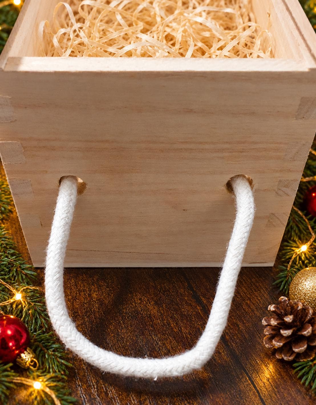 Snowman wine bottle carrier on a wooden table with red and gold baubles, evergreen sprigs and a lit Christmas tree in the background.