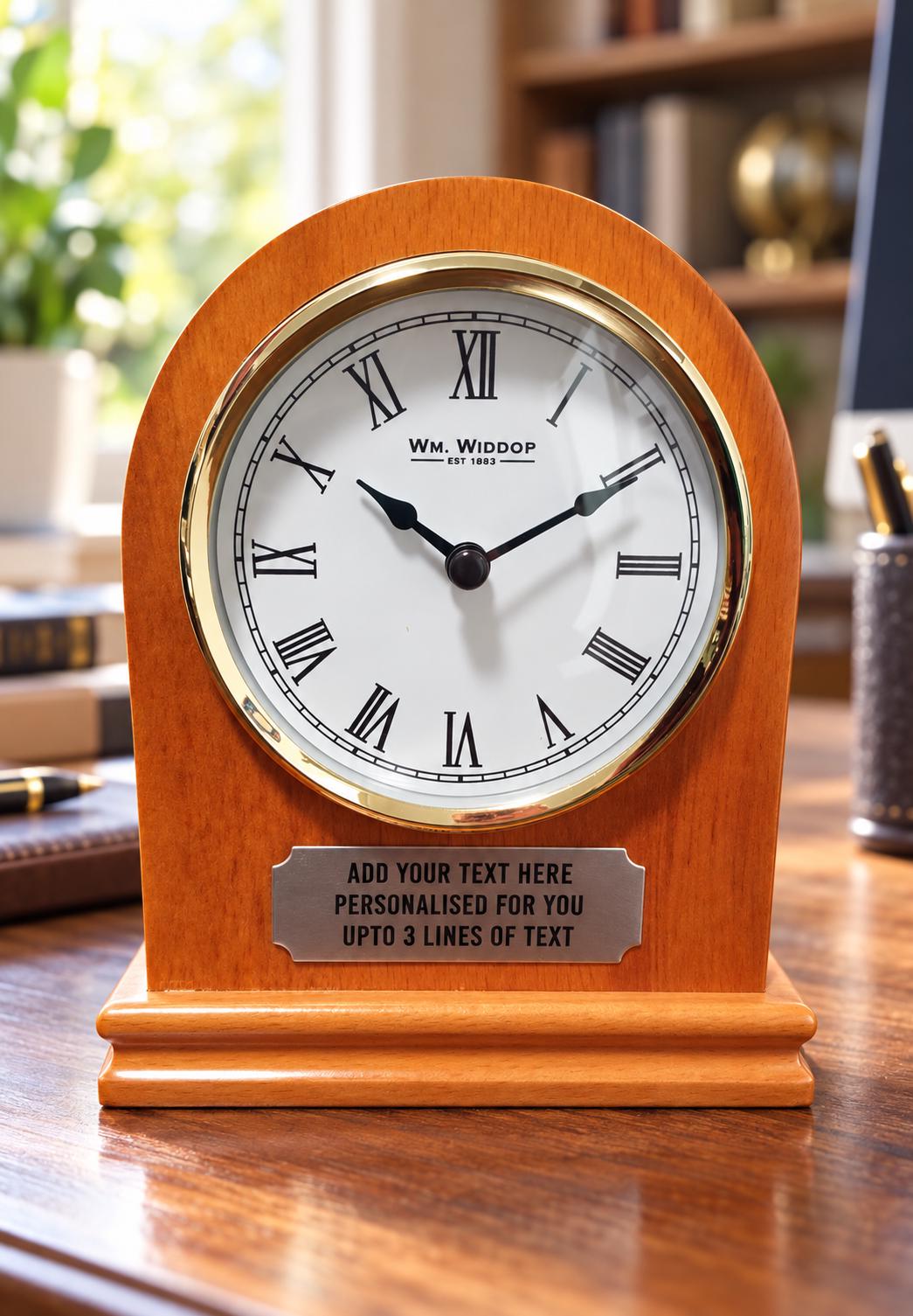 Personalised wooden arched mantel clock with a Roman numeral dial, gold-tone surround and engraved plaque on a home office desk with a blurred background.