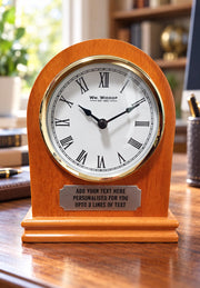 Personalised wooden arched mantel clock with a Roman numeral dial, gold-tone surround and engraved plaque on a home office desk with a blurred background.
