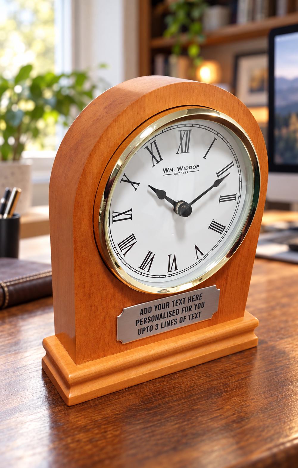 Angled view of a wooden arched desk clock showing its stepped base, Roman numeral face and three-line personalised plaque against a softly blurred home office.