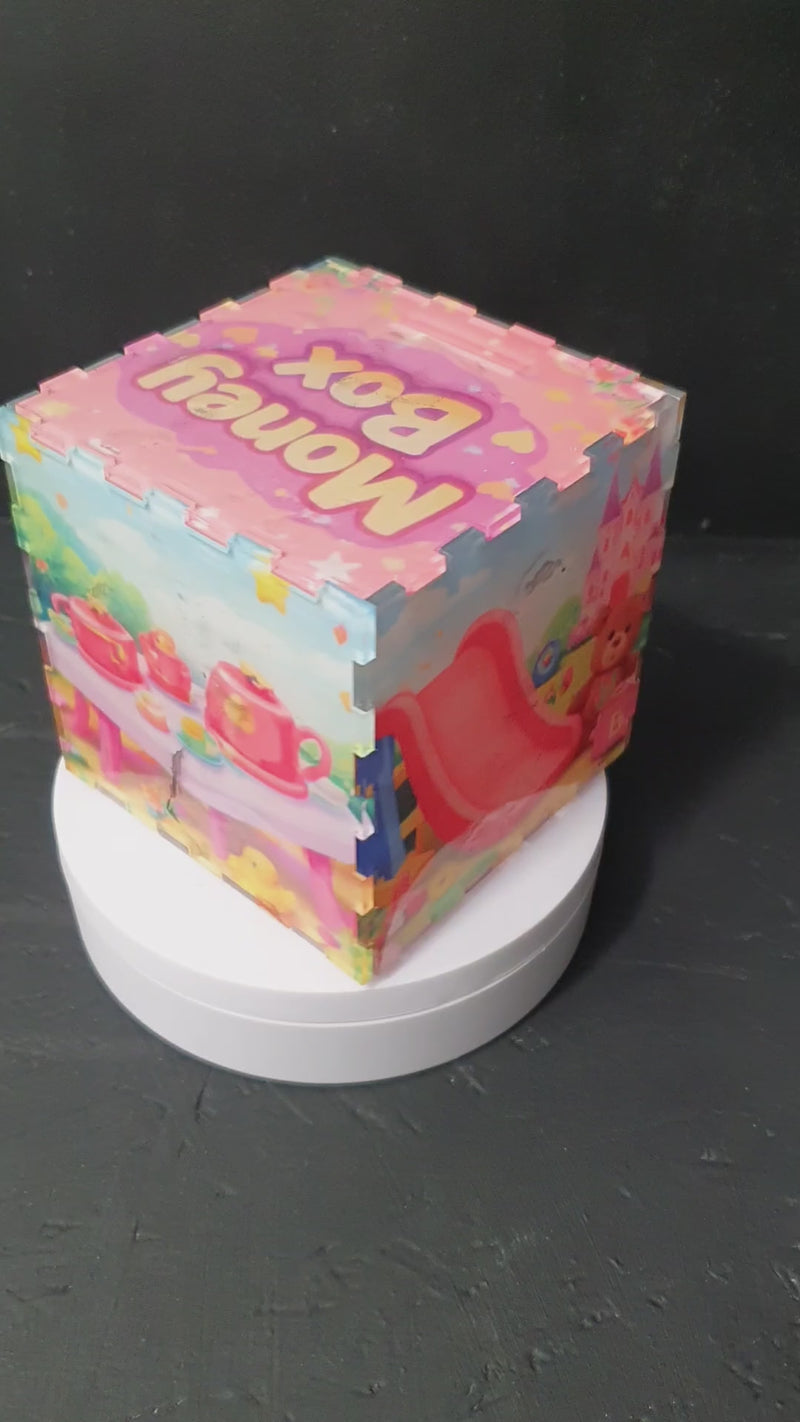 Front view of pink children’s money box with “Money Box” text on lid and pastel toy-themed design, 10x10x10cm acrylic piggy bank.