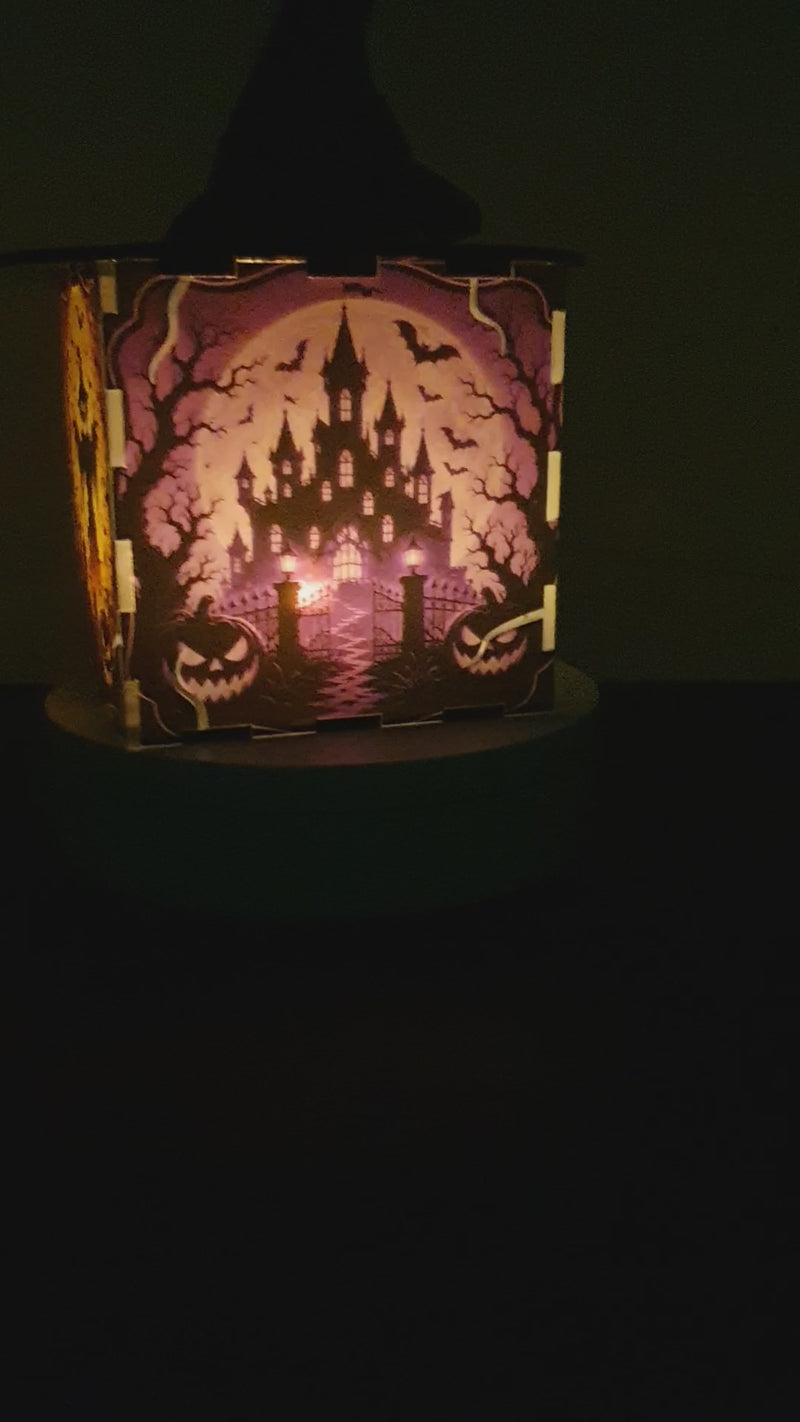 A Halloween-themed decorative box with a black witch hat on top and a spooky orange and black haunted-house illustration on the front, displayed on a white pedestal against a white wall.