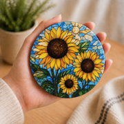 A hand holds a round pinback button featuring a stained-glass style sunflower design with yellow petals, dark brown centers, and green leaves against a blue background.